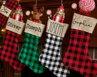 May include: Four Christmas stockings hang from a mantel, each with a different plaid pattern: red, green, and black and white. Each stocking is personalized with a name: Star, Oliver, Joseph, and Sophia. Decorations include a paw print, nutcrackers, a reindeer, and a candy cane.