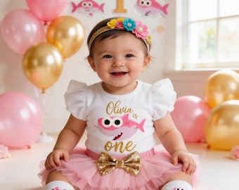 May include: A baby girl wearing a white shirt with a pink shark graphic and the text "Olivia is one." She is wearing a pink tutu skirt, a gold bow, and a floral headband. Pink and gold balloons are in the background.