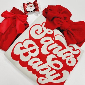 May include: A festive ensemble featuring a white shirt with the text "Santa Baby" in red, accompanied by a red bow, a red lace bow, and a Santa Claus bow. The items are arranged on a white surface.