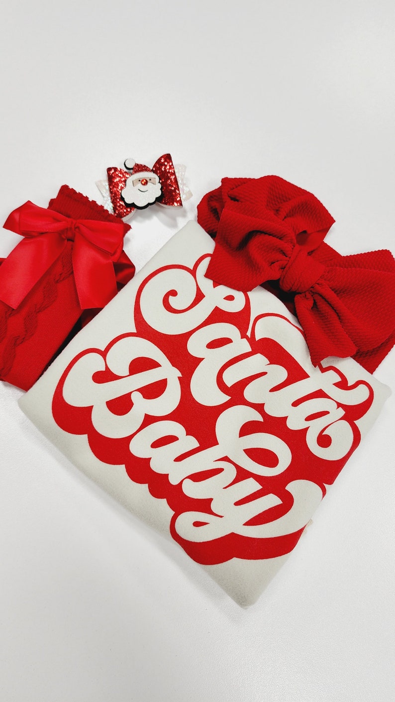 May include: A festive ensemble featuring a white shirt with the text "Santa Baby" in red, accompanied by a red bow, a red lace bow, and a Santa Claus bow. The items are arranged on a white surface.
