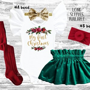May include: A white bodysuit with the text "My First Christmas" in gold glitter, surrounded by a wreath of red roses and green leaves. The bodysuit has ruffled sleeves and is paired with a green velvet skirt and red leg warmers. Two headbands are included, one gold glitter bow and one red.