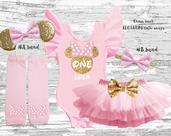 May include: Pink and gold Minnie Mouse themed first birthday outfit for a baby girl. The outfit includes a pink leotard with gold glitter, a pink tulle skirt with a gold bow, pink leg warmers with white polka dots, a pink headband with gold glitter, and a gold headband with pink ears and a bow.
