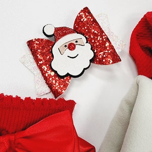 May include: A festive Christmas hair bow featuring a Santa Claus face in the center. The bow has red glitter accents and white details. Other red and white Christmas-themed accessories are also visible.