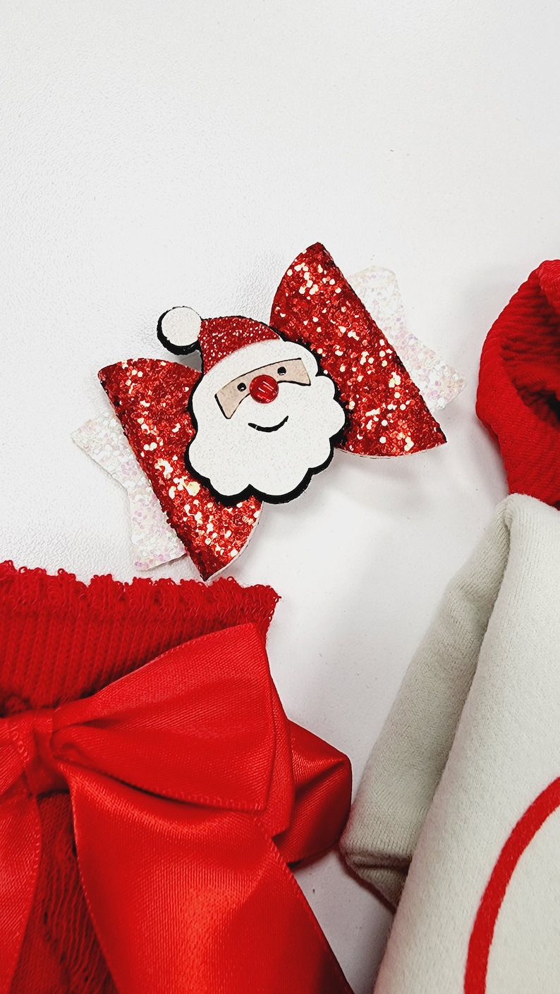 May include: A festive Christmas hair bow featuring a Santa Claus face in the center. The bow has red glitter accents and white details. Other red and white Christmas-themed accessories are also visible.