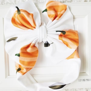 May include: White headband with a large bow and a pumpkin print. The headband has the text "Lily and Max Co." printed on the bottom.