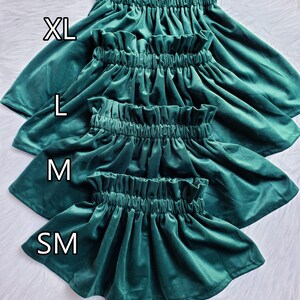Four dark green velvet skirts stacked vertically on a white surface, labeled with sizes XL, L, M, and SM, with text at the bottom stating that X-SM size is coming soon.