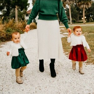 May include: A woman in a green sweater and white skirt walks with two young girls. The girls are wearing white sweaters and matching skirts, one green and one red.
