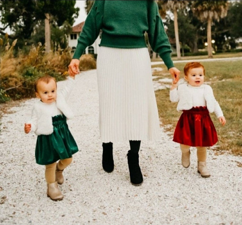 May include: A woman in a green sweater and white skirt walks with two young girls. The girls are wearing white sweaters and matching skirts, one green and one red.