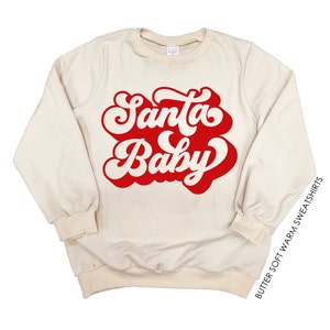 May include: Cream-colored sweatshirt with the words "Santa Baby" in a red, retro font. The long-sleeved top has a crew neck and ribbed cuffs and hem. The text "BUTTER SOFT WARM SWEATSHIRTS" is printed vertically on the right side.