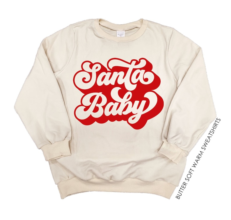 May include: Cream-colored sweatshirt with the words "Santa Baby" in a red, retro font. The long-sleeved top has a crew neck and ribbed cuffs and hem. The text "BUTTER SOFT WARM SWEATSHIRTS" is printed vertically on the right side.