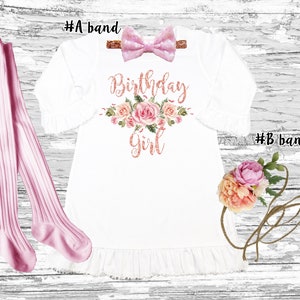 K&ouml;nnte beinhalten: Ein wei&szlig;es Kleid mit einem rosafarbenen Blumendesign und dem Text "Birthday Girl" in ros&eacute;goldenen Glitzer. Das Kleid hat einen R&uuml;schen-Saum und wird mit einem rosafarbenen Schleifen-Haarreif und rosafarbenen Strumpfhosen geliefert.