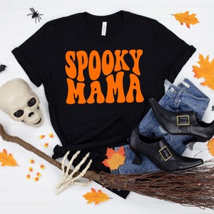 May include: Black t-shirt with the text "Spooky Mama" in orange retro font. The shirt is surrounded by Halloween decorations, including a witch's broom, a skull, a skeleton hand, and fall leaves.