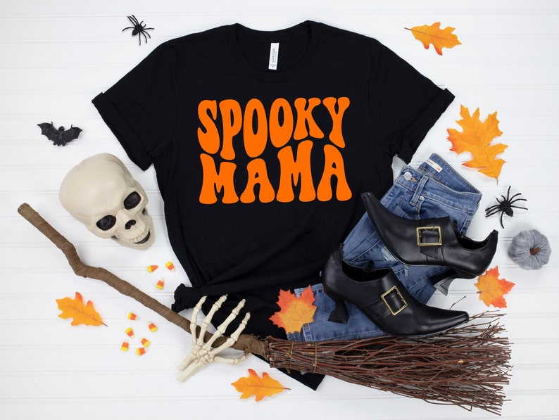 May include: Black t-shirt with the text "Spooky Mama" in orange retro font. The shirt is surrounded by Halloween decorations, including a witch's broom, a skull, a skeleton hand, and fall leaves.