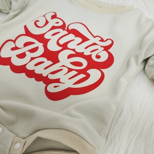 May include: A light green baby onesie with the words "Santa Baby" in red script. The onesie has beige trim around the neckline, sleeves, and legs. It features snap closures and long sleeves.