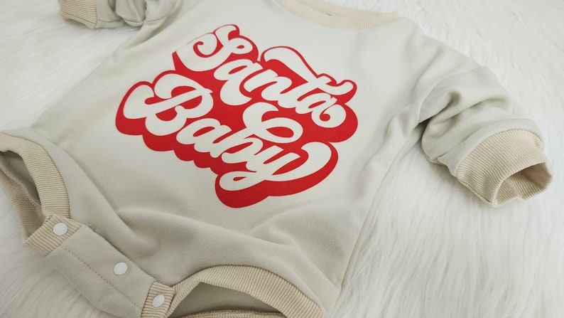 May include: A light green baby onesie with the words "Santa Baby" in red script. The onesie has beige trim around the neckline, sleeves, and legs. It features snap closures and long sleeves.