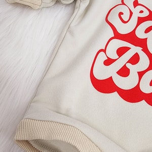 May include: Cream-colored baby onesie with red text. The onesie has tan ribbed cuffs and trim. The text is in a bold, retro font. The onesie is made of a soft material and has snap closures.
