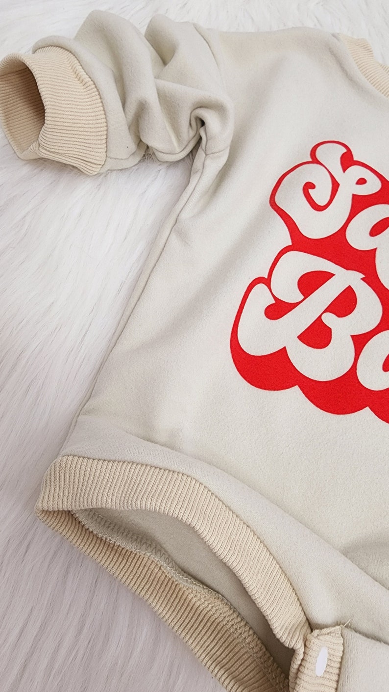 May include: Cream-colored baby onesie with red text. The onesie has tan ribbed cuffs and trim. The text is in a bold, retro font. The onesie is made of a soft material and has snap closures.