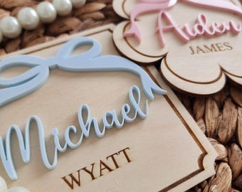 May include: Two wooden name plaques with a bow and name in raised lettering. The plaque on the left is light blue with the name "Michael Wyatt" and the plaque on the right is pink with the name "Arden James".
