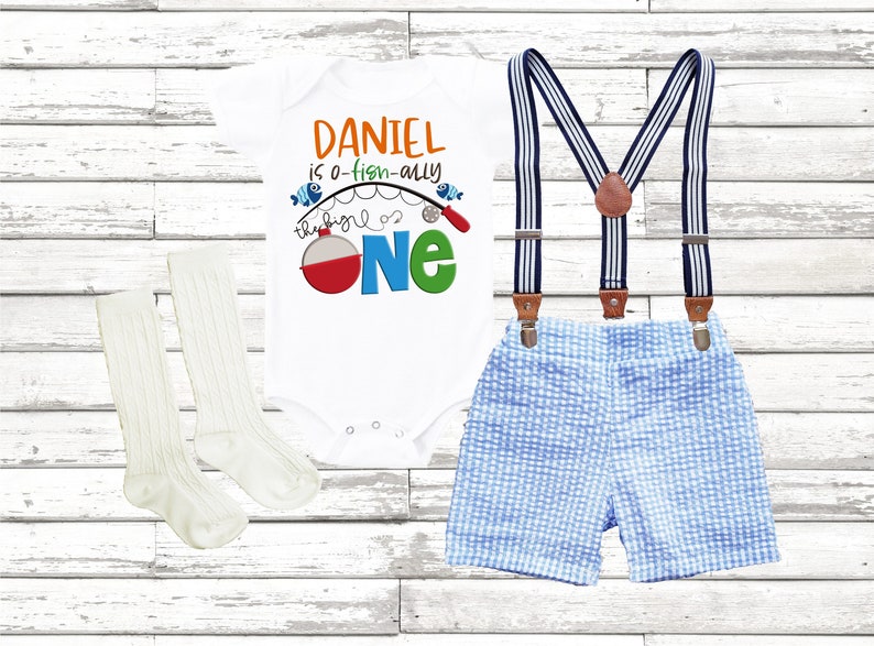 Puede incluir: Un body blanco con el texto "Daniel is a-fish-ally the big one" y una ca&ntilde;a de pescar con un flotador rojo. El body se combina con pantalones cortos de cuadros azules y blancos y calcetines blancos.