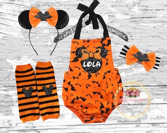 Puede incluir: Conjunto de disfraz de Halloween naranja y negro para bebé. El conjunto incluye un body con estampado de murciélagos negro y naranja, una diadema con orejas negras y un lazo, y polainas con rayas negras y naranjas. El body tiene una cabeza de Mickey Mouse negra con purpurina con el nombre "Lola" en ella.