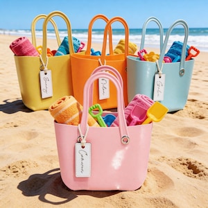 May include: Five brightly coloured beach bags filled with towels and toys on a sandy beach. The bags are yellow, orange, pink, blue, and light blue. Each bag has a white tag with a name. The bags have curved handles.