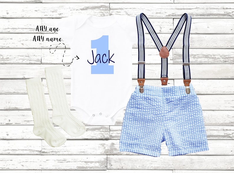 May include: A white bodysuit with a blue number one and the name "Jack" printed on it. The bodysuit is paired with blue and white checkered shorts and white suspenders. The outfit is completed with a pair of white knee-high socks.