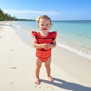 May include: A smiling toddler wearing a red and black patterned swimsuit with ruffled sleeves stands on a sandy beach. A white flower adorns the child's hair. The background features a clear blue sky and turquoise water.
