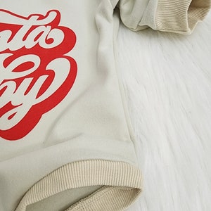 May include: Cream-colored baby bodysuit with the text "Santa Baby" in red script. The long-sleeved onesie has ribbed cuffs and trim in a tan color. The garment is made of a soft material and has a snap closure.