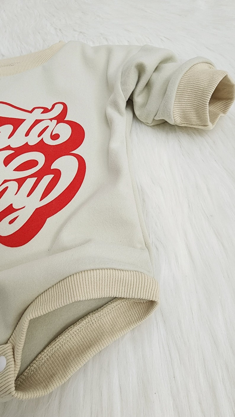 May include: Cream-colored baby bodysuit with the text "Santa Baby" in red script. The long-sleeved onesie has ribbed cuffs and trim in a tan color. The garment is made of a soft material and has a snap closure.