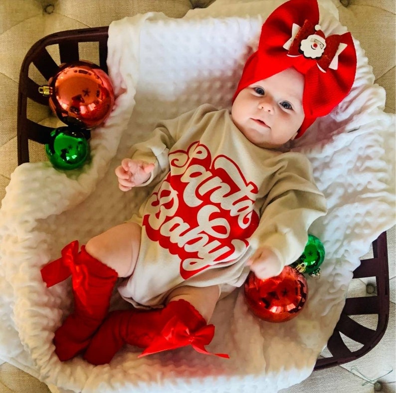May include: A baby wearing a tan long-sleeved bodysuit with the words 'Santa Baby' in red letters. The baby is wearing a red bow headband and red leg warmers with bows. The baby is lying on a white blanket with red and green Christmas ornaments.