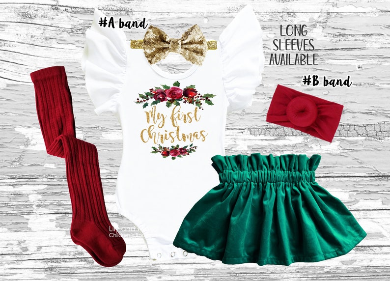 May include: A white bodysuit with the text "My First Christmas" in gold glitter, surrounded by a wreath of red roses and green leaves. The bodysuit has ruffled sleeves and is paired with a green velvet skirt and red knit leggings. Two headbands are included, one gold glitter bow and one red.