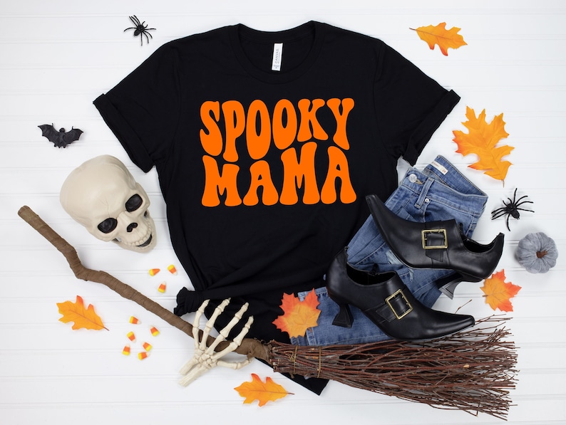 May include: Black t-shirt with the text "Spooky Mama" in orange retro font. The shirt is laid out on a white surface with a witch's broom, a skull, and fall leaves.