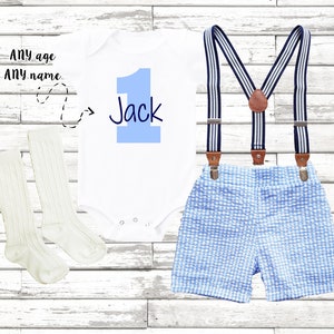May include: A white onesie with a blue number one and the name Jack written in black. The onesie is paired with blue and white gingham shorts and suspenders. The outfit is completed with a pair of white socks.