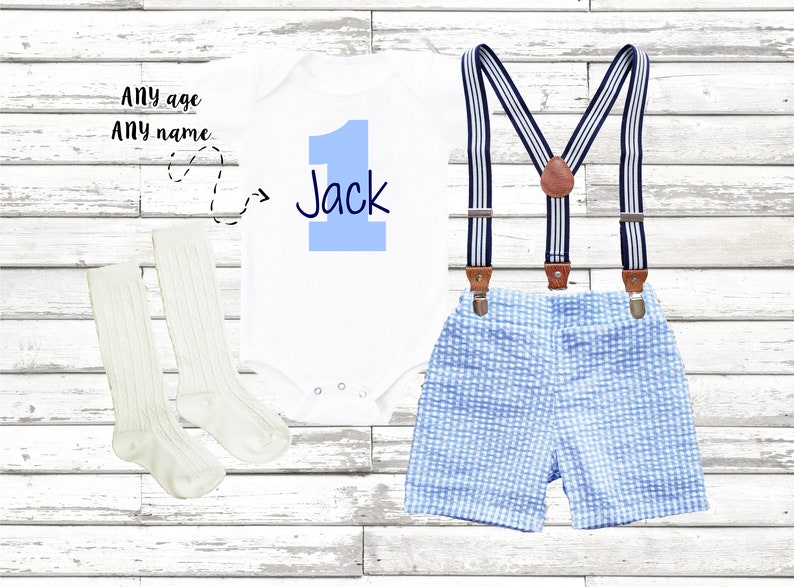 May include: A white onesie with a blue number one and the name Jack written in black. The onesie is paired with blue and white gingham shorts and suspenders. The outfit is completed with a pair of white socks.