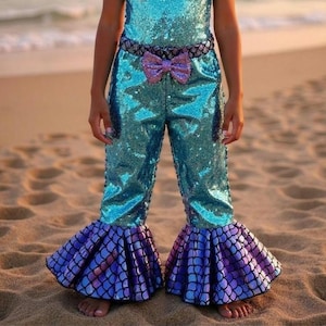 May include: A turquoise sequined jumpsuit with a mermaid theme. The pants have flared bottoms in a scale pattern of purple and blue. A purple bow is on the front, and a scale-patterned belt is at the waist.
