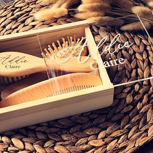 May include: A light-colored wooden box with a clear acrylic sliding lid featuring the white text Addie Claire. Inside the box are a wooden hairbrush and a wooden comb, both engraved with the name Addie Claire in a black cursive script font.
