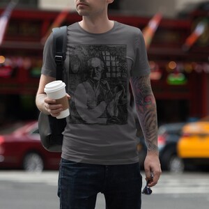 May include: A man wearing a gray t-shirt with a black and white graphic print of a man holding a glass. The graphic print is of a man with a mustache and beard. The man is holding a cup of coffee in his left hand.