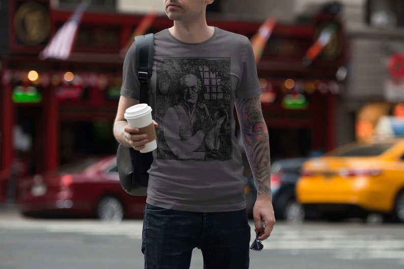 May include: A man wearing a gray t-shirt with a black and white graphic print of a man holding a glass. The graphic print is of a man with a mustache and beard. The man is holding a cup of coffee in his left hand.