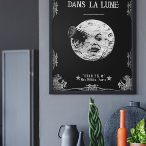 Puede incluir: Un p&oacute;ster en blanco y negro con un dise&ntilde;o vintage que presenta una luna con una cara y un telescopio. El texto en el p&oacute;ster dice "Le Voyage Dans La Lune" y "Star Film Geo M&eacute;li&egrave;s Paris".
