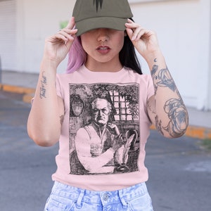 May include: A woman wearing a pink t-shirt with a black and white illustration of a man with a quill pen. She is also wearing a green baseball cap with a black logo and blue jeans.