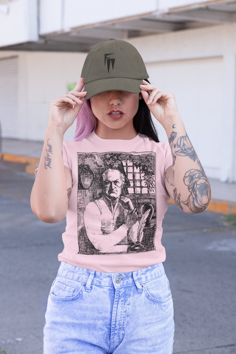 May include: A woman wearing a pink t-shirt with a black and white illustration of a man with a quill pen. She is also wearing a green baseball cap with a black logo and blue jeans.