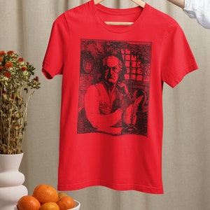 May include: Red t-shirt with a black and white illustration of a man sitting at a desk with a quill pen and inkwell. The illustration is surrounded by a black border. The text "I'll Come Back For The Rest" is visible in the illustration.