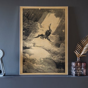 Puede incluir: Impresi&oacute;n vintage enmarcada que representa una figura alada cayendo a trav&eacute;s de las nubes, iluminada por un rayo de luz. La obra de arte es en blanco y negro con un tono sepia, sobre una pared gris oscuro. Un reloj decorativo, una hoja dorada y un tel&eacute;fono rosa est&aacute;n en el estante.