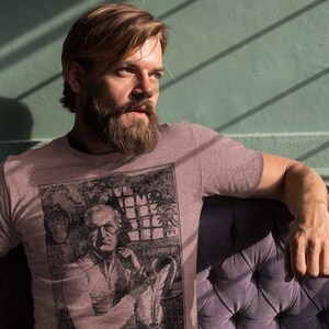 May include: A heather grey t-shirt with a vintage illustration of a man with a beard and mustache. The illustration is in black and white and features a man sitting in a chair with a book in his hand.