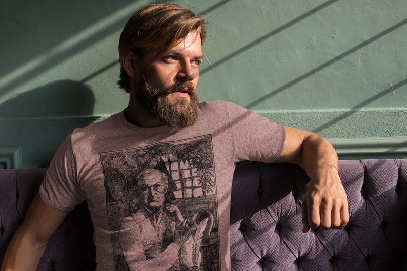 May include: A heather grey t-shirt with a vintage illustration of a man with a beard and mustache. The illustration is in black and white and features a man sitting in a chair with a book in his hand.