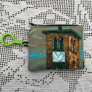May include: A small, rectangular coin purse with a green zipper and a green clip. The purse features a printed design of a building with a clock tower in shades of orange, green, and blue.