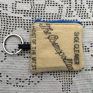 May include: A small, rectangular, beige shoe cleaner pouch with a blue zipper and a silver keyring. The pouch has black text that reads "The Goodfoot" and "Shoe Cleaner". The text "Host of the West" is also printed on the side.