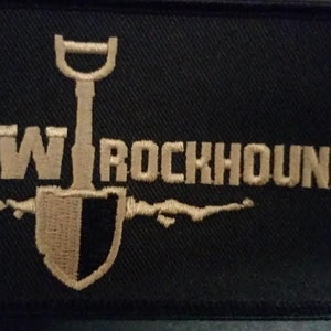 May include: A black embroidered patch with the text "NW ROCKHOUNDS" in beige thread. A beige shovel with a black handle is centered above the text.