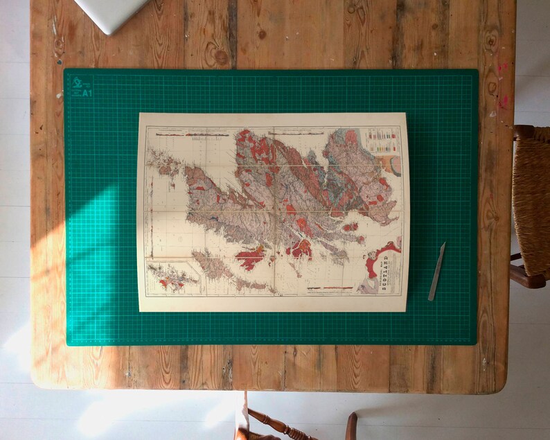 May include: A vintage map of the British Isles, printed on paper, is laid out on a green cutting mat. The map is surrounded by a wooden table surface.