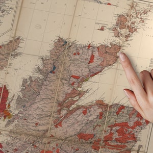 May include: A vintage map of Scotland with a finger pointing to the location of the Isle of Skye. The map is printed on paper with a cream background and features red, blue, and brown lines and markings.